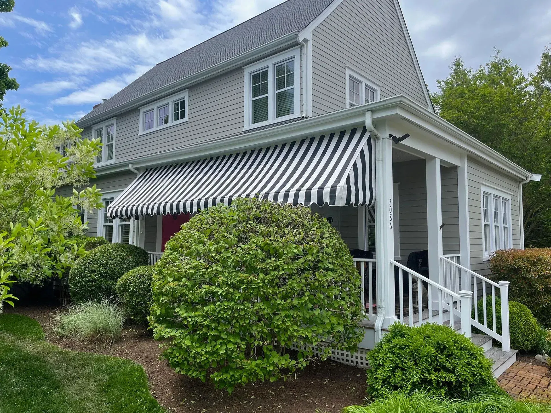 A two-story grey shingled house with white trim, a black and white striped awning, and manicured green bushes in front.