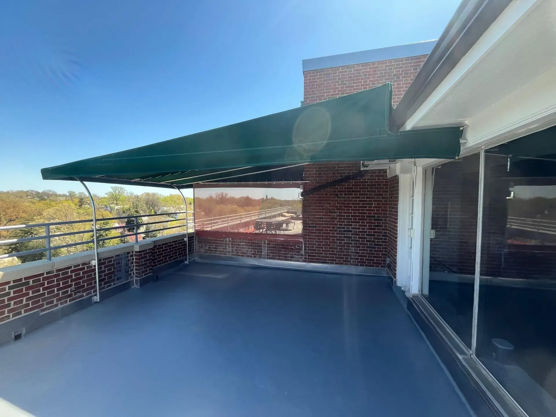 An outdoor balcony with a dark green retractable awning, gray flooring, brick walls, and a metal railing.