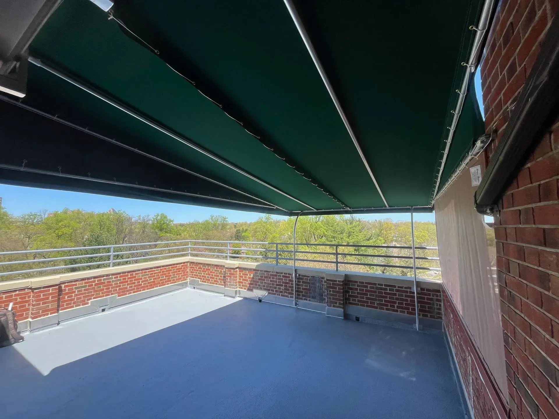 A rooftop patio with a gray floor, brick walls, a metal railing, and a large green retractable awning under a blue sky.