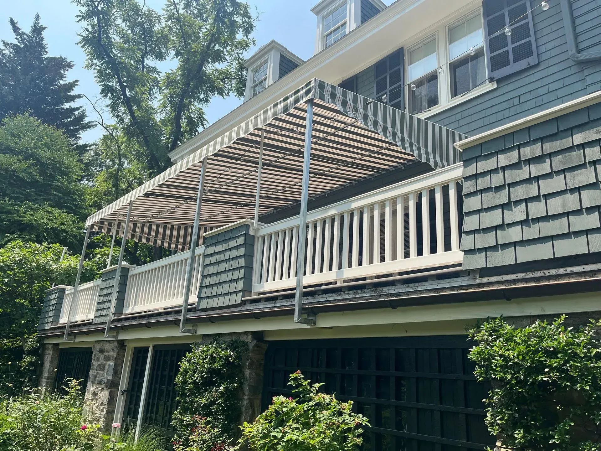 A blue-gray house with a second-story balcony featuring a striped fabric awning and white railing, surrounded by trees.