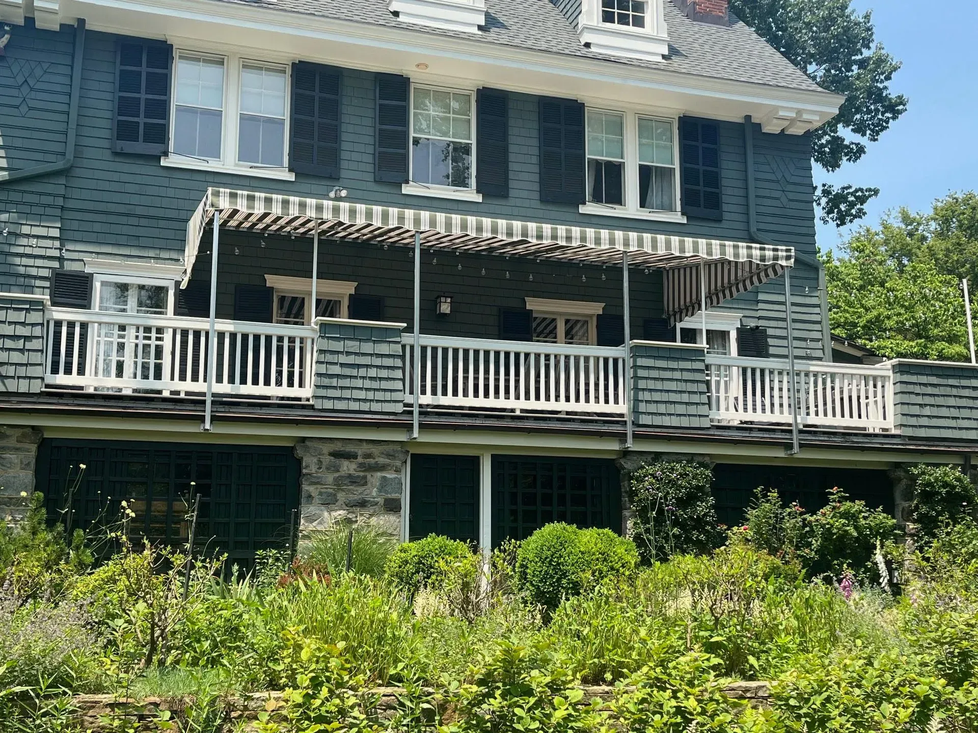 A dark gray two-story house with a white railed balcony, striped awning, and stone foundation behind dense green bushes.