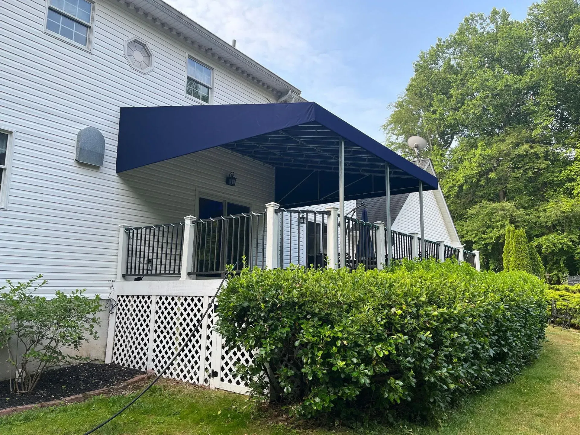 A dark blue awning covers a white residential deck with black metal railings, lattice skirting, and surrounding bushes.