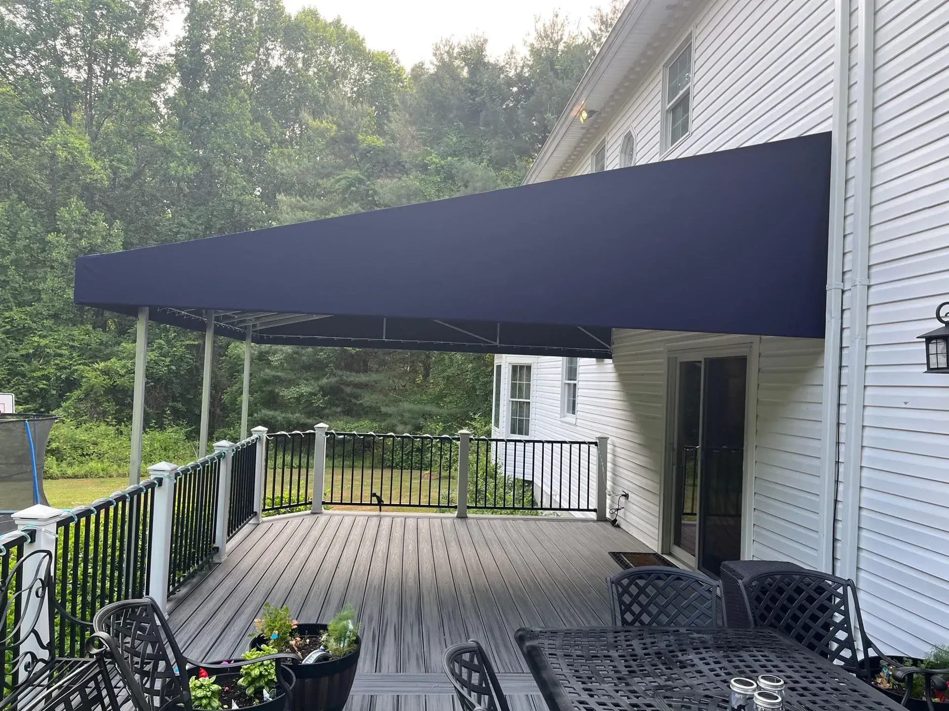 A large, dark blue rectangular awning extends from a white-sided house over a wooden deck with outdoor furniture.