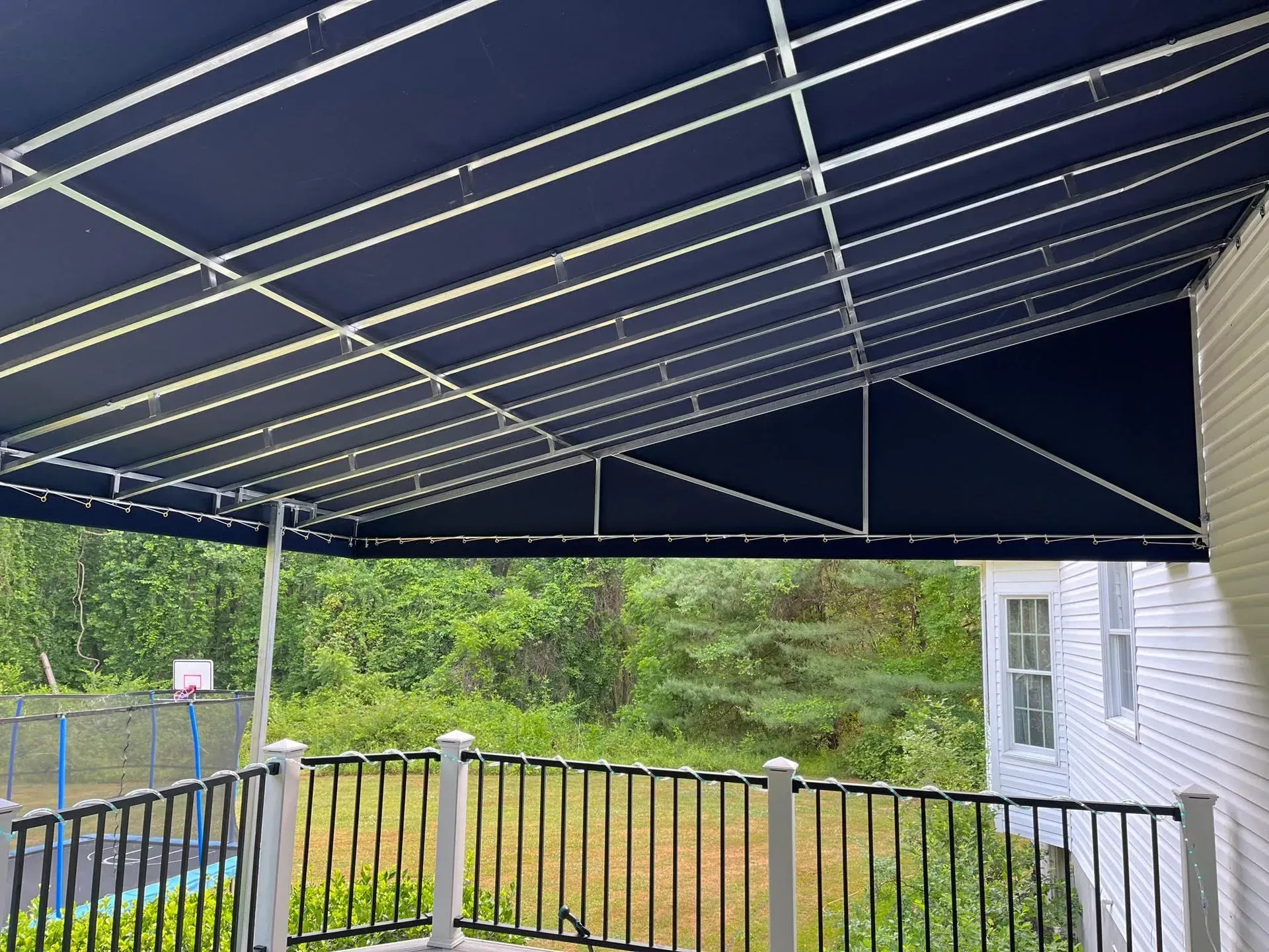 A dark blue fabric awning covers a deck with a black railing, overlooking a green, wooded backyard.