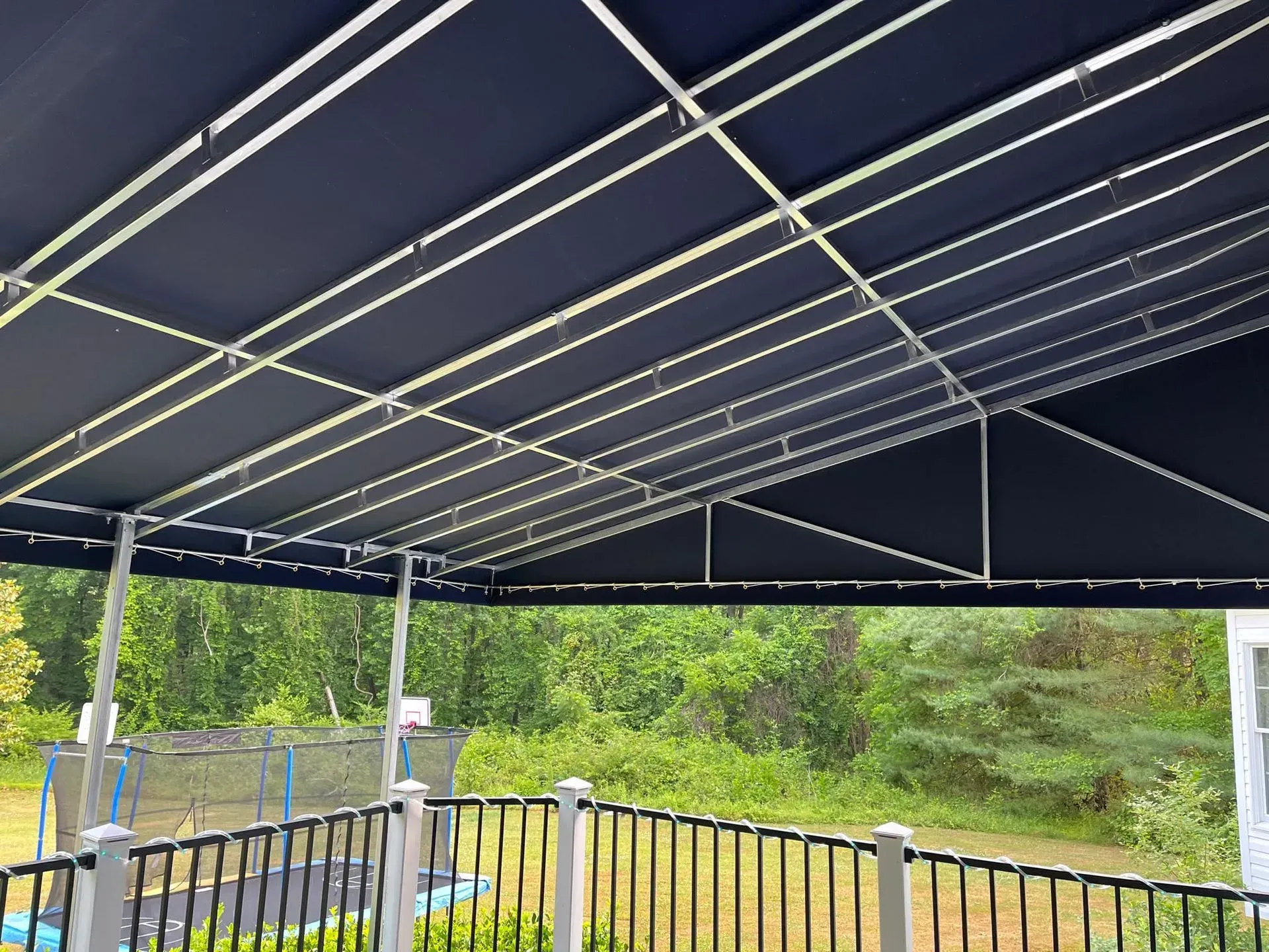 A dark blue metal-framed canopy over a deck railing, overlooking a green backyard with a trampoline in the distance.