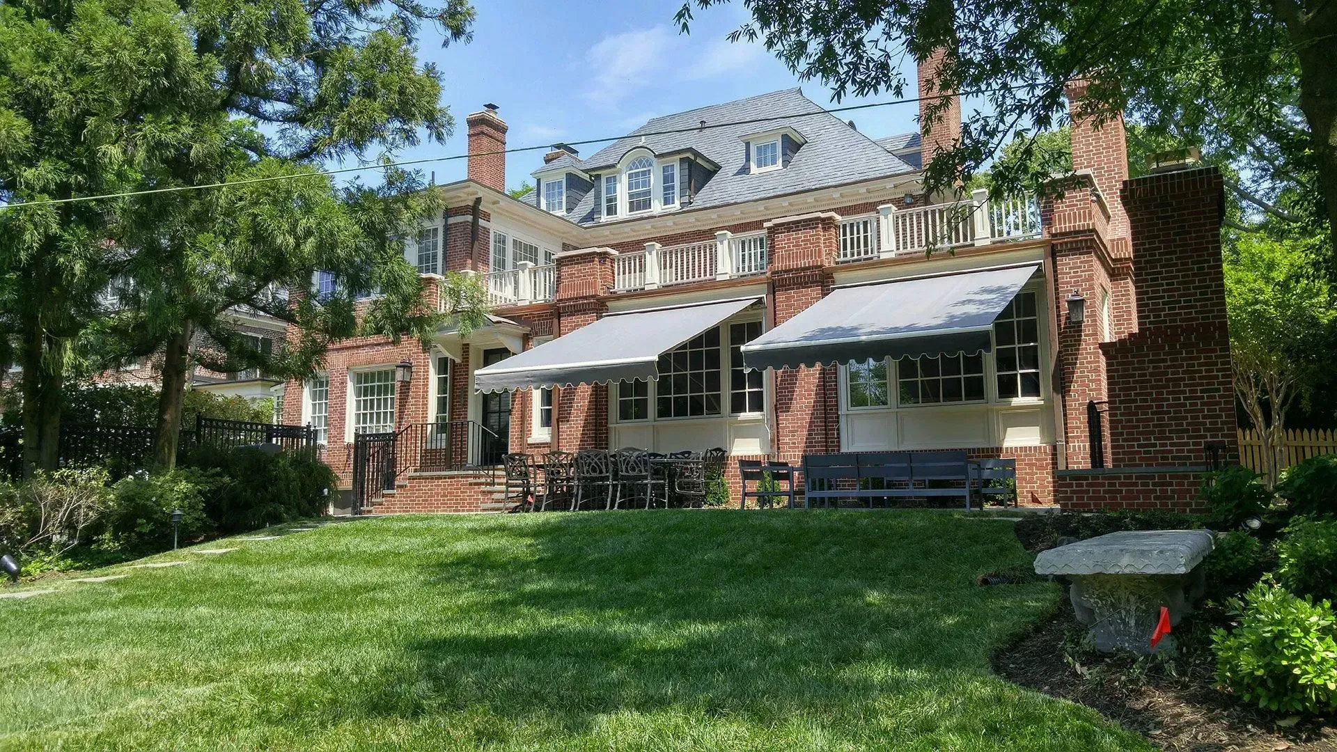 A two-story brick mansion with dormer windows, a slate roof, and two gray retractable awnings overlooking a green lawn.