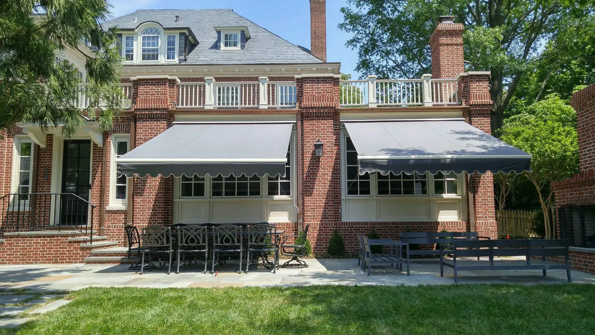 A brick house with two gray retractable awnings shading a stone patio featuring outdoor dining sets and a lawn.