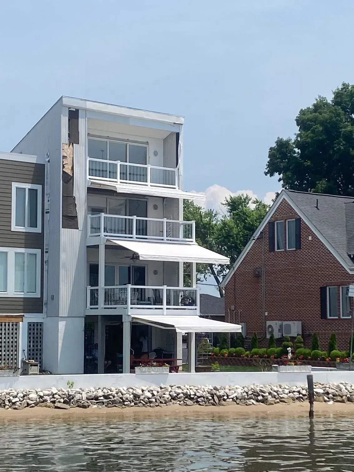 A modern, three-story white residential building with retractable awnings on each balcony, situated on a waterfront lot.