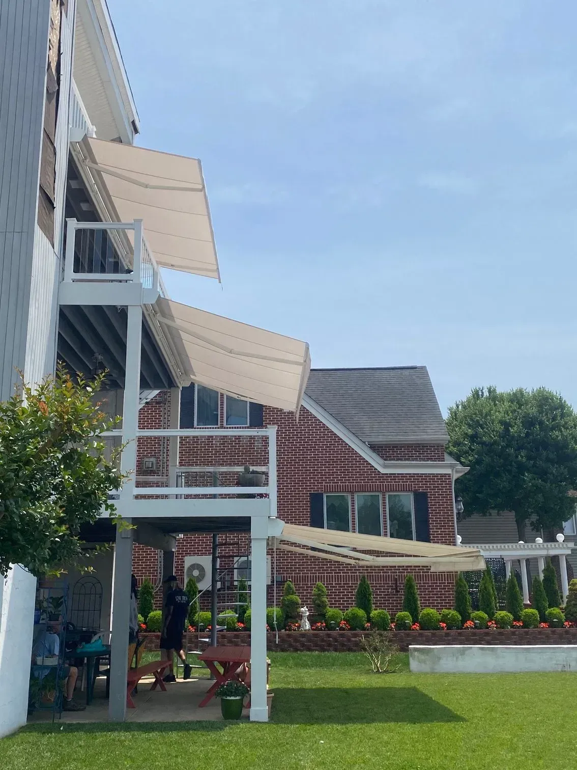 A side view of a multi-story white deck with beige awnings, set against a red brick house and a green lawn.