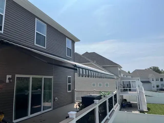 Dark-sided house with a white railing deck and a partially extended striped retractable awning over a sliding glass door.