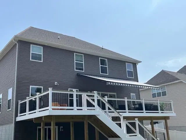 A two-story house with dark grey siding and a large white deck with a retractable awning and stairs.