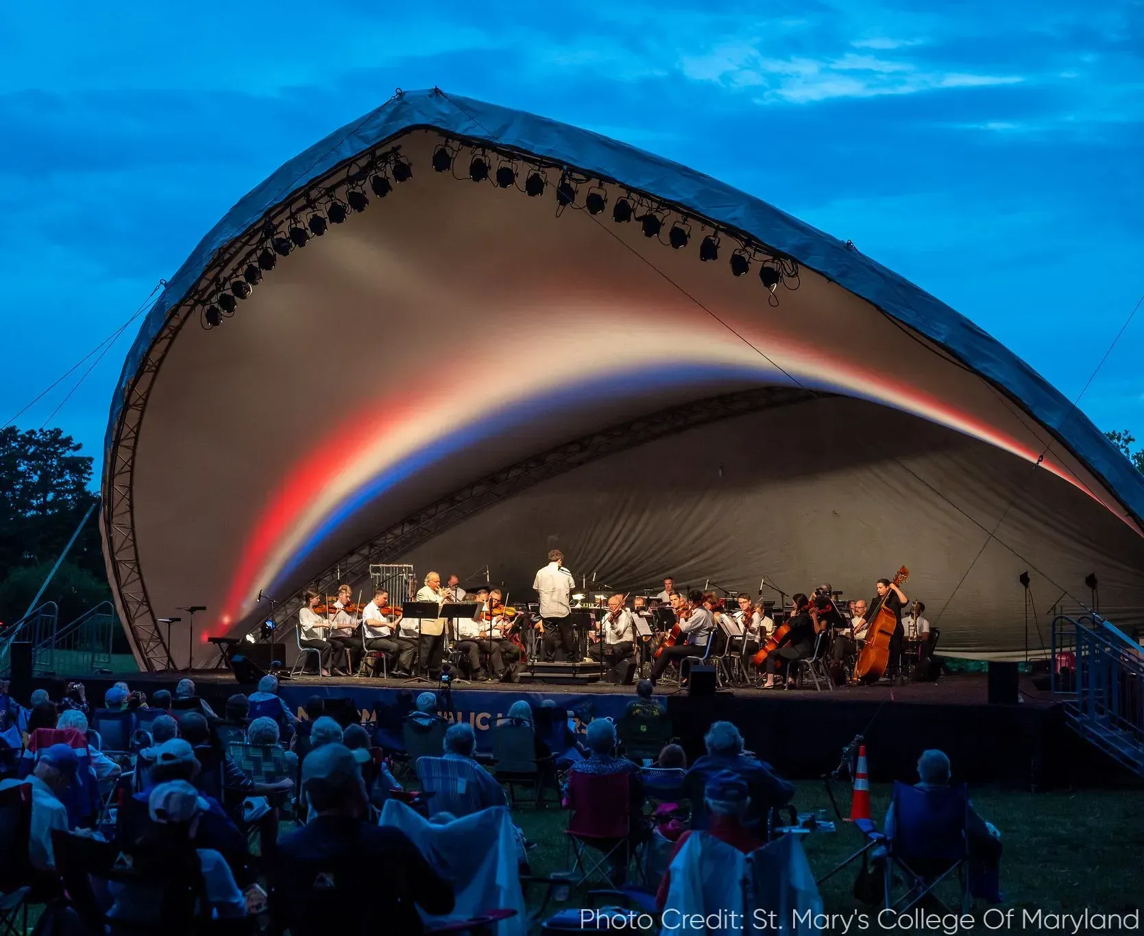 An orchestra performs on a stage under a large, illuminated acoustic shell at twilight, viewed by an outdoor audience.
