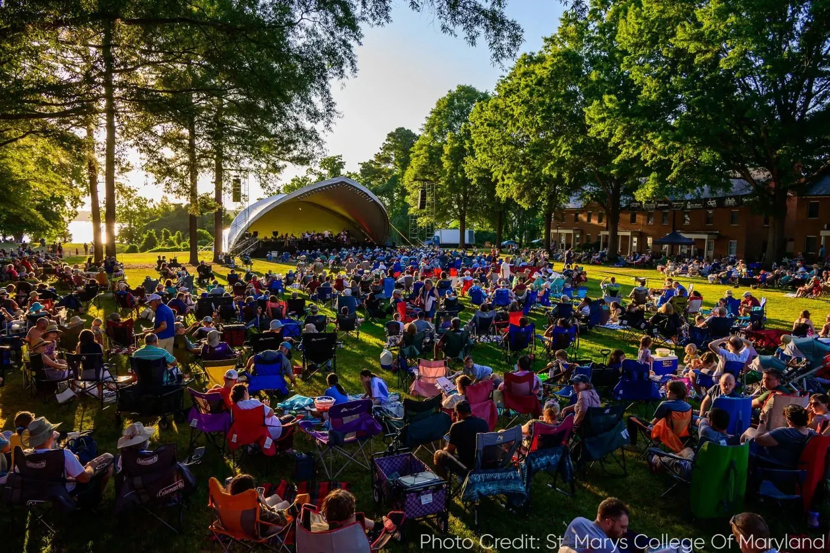 A large crowd sits in folding chairs and on blankets on a grassy lawn facing an outdoor bandshell at sunset.