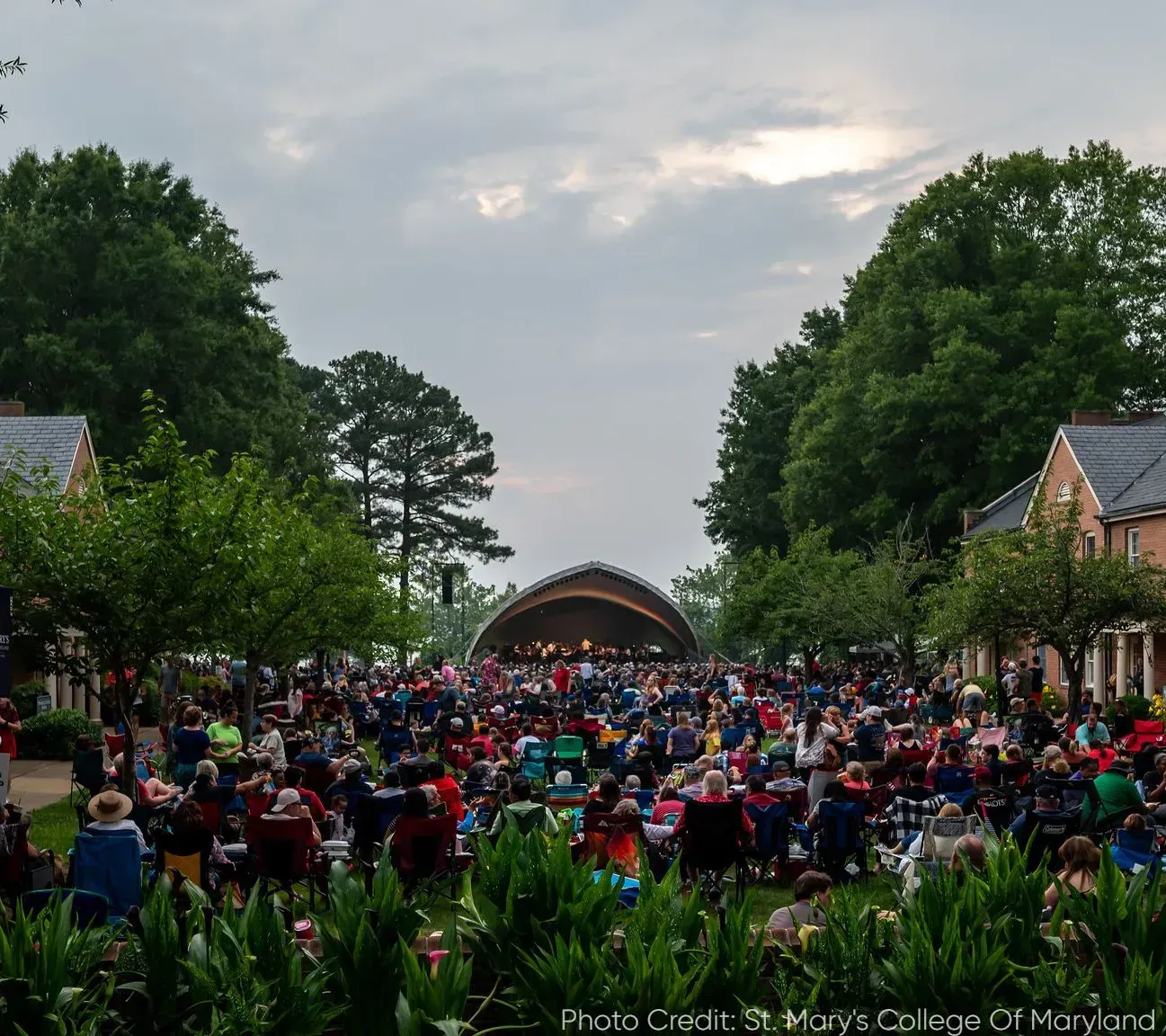 A large crowd gathered in a grassy outdoor amphitheater to watch a concert under a domed stage at twilight.