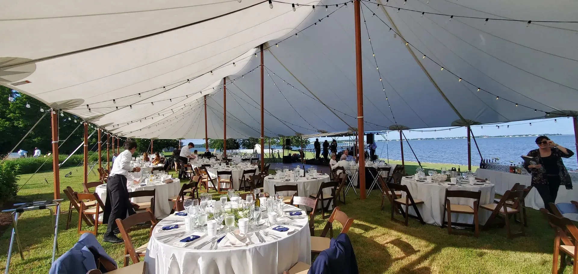 A white tent on a lawn by the water, set up for an event with round tables featuring white tablecloths and place settings.