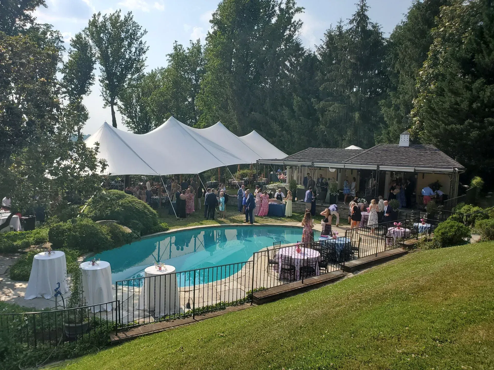 Outdoor party with a large white tent, swimming pool, and round tables set on a lawn near a covered patio and trees.