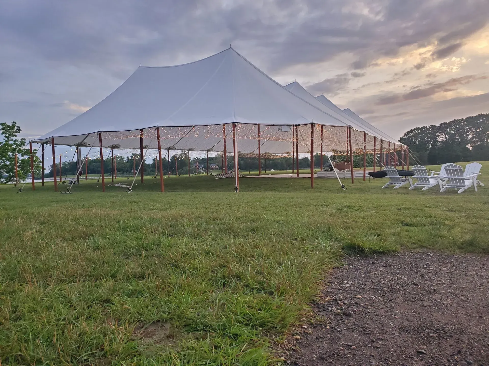 A large white event tent with string lights stands on a grassy field under a cloudy sky at sunset.