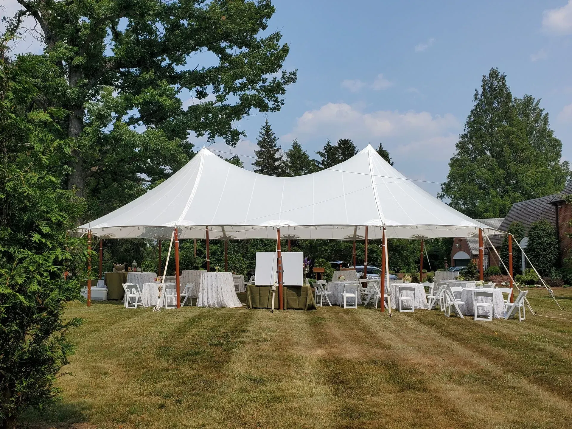 A large white event tent set up on a grassy lawn with tables and chairs for an outdoor gathering under a blue sky.