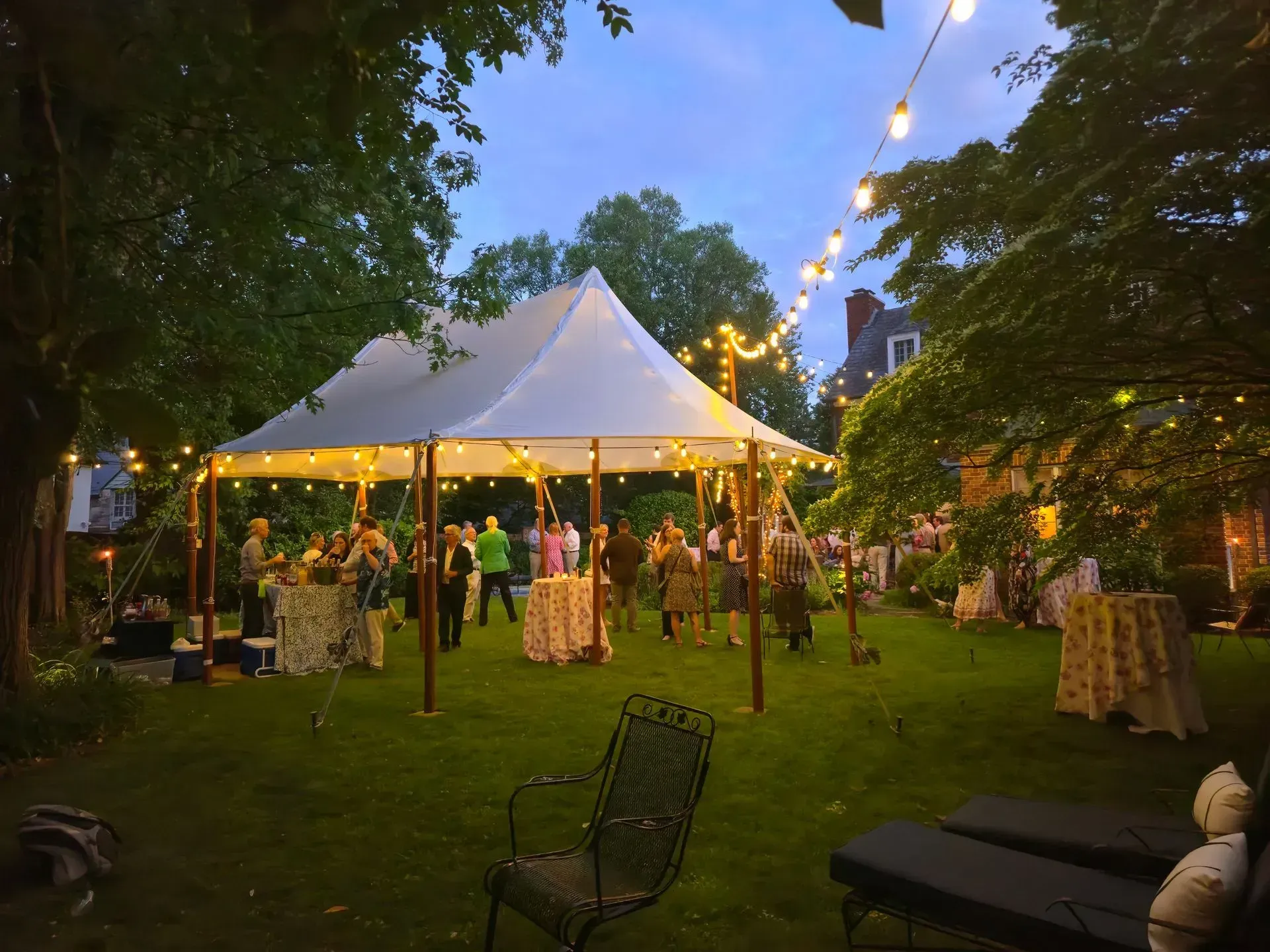 People mingle under a white event tent in a lush, green backyard at dusk, illuminated by glowing string lights.