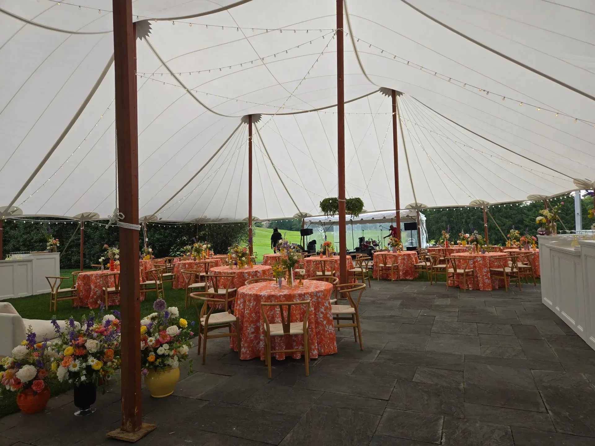 A white tent hosts an event with orange-patterned circular tables, wooden chairs, and floral arrangements on a stone patio.