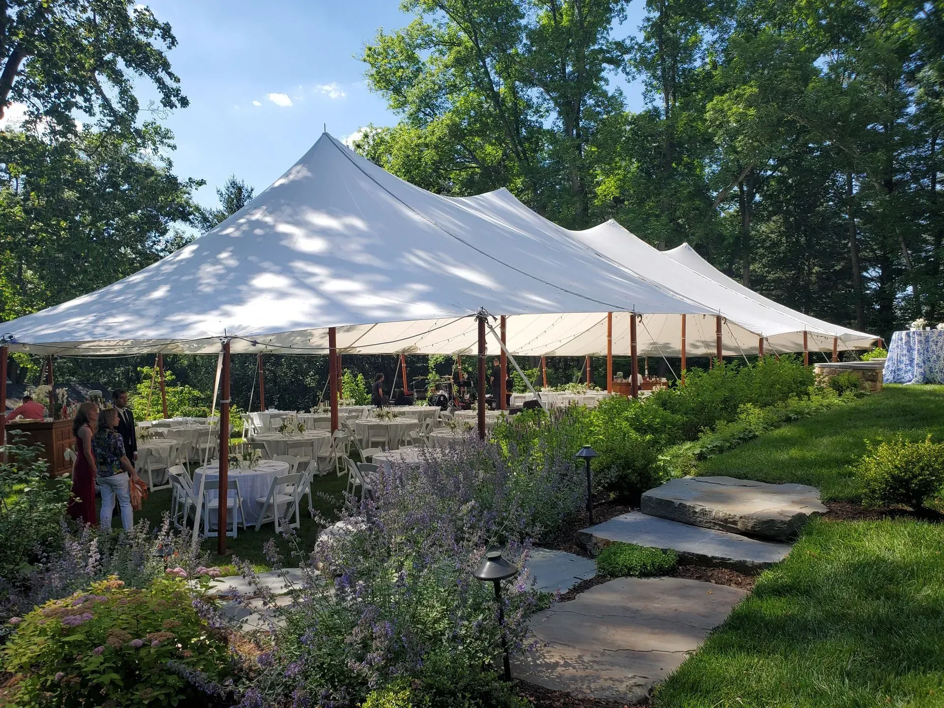 A large white party tent set up on a lawn with tables, chairs, and guests under the canopy on a sunny day.