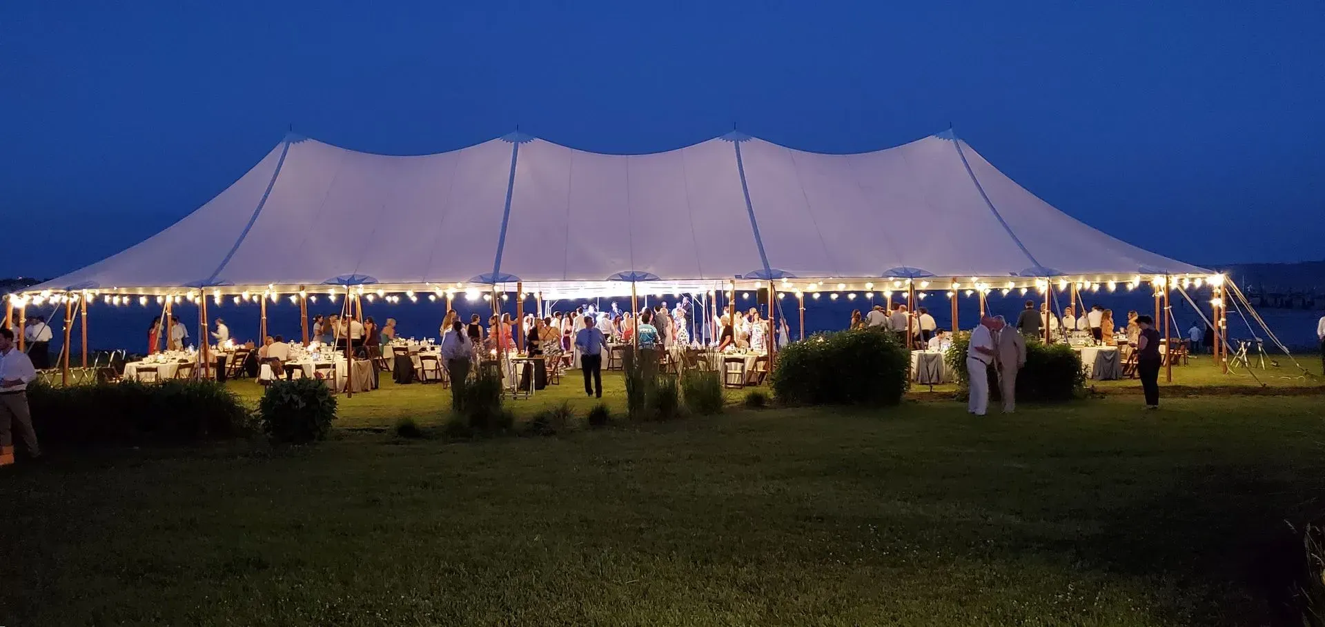 A large white event tent lit by string lights at dusk, with guests dining inside on a grassy lawn near the water.