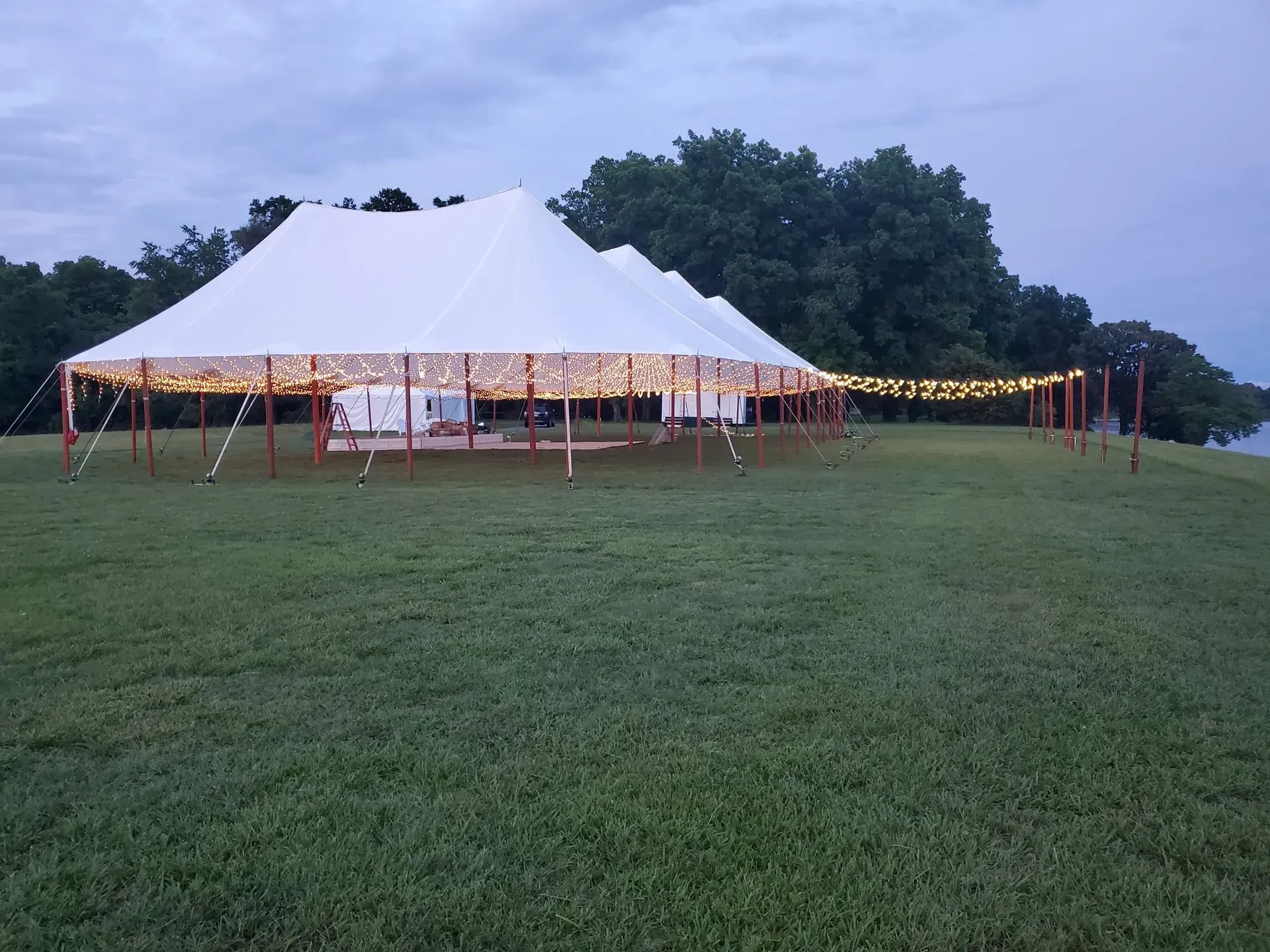 A large white event tent lit with string lights stands on a grassy field at twilight, with trees in the background.