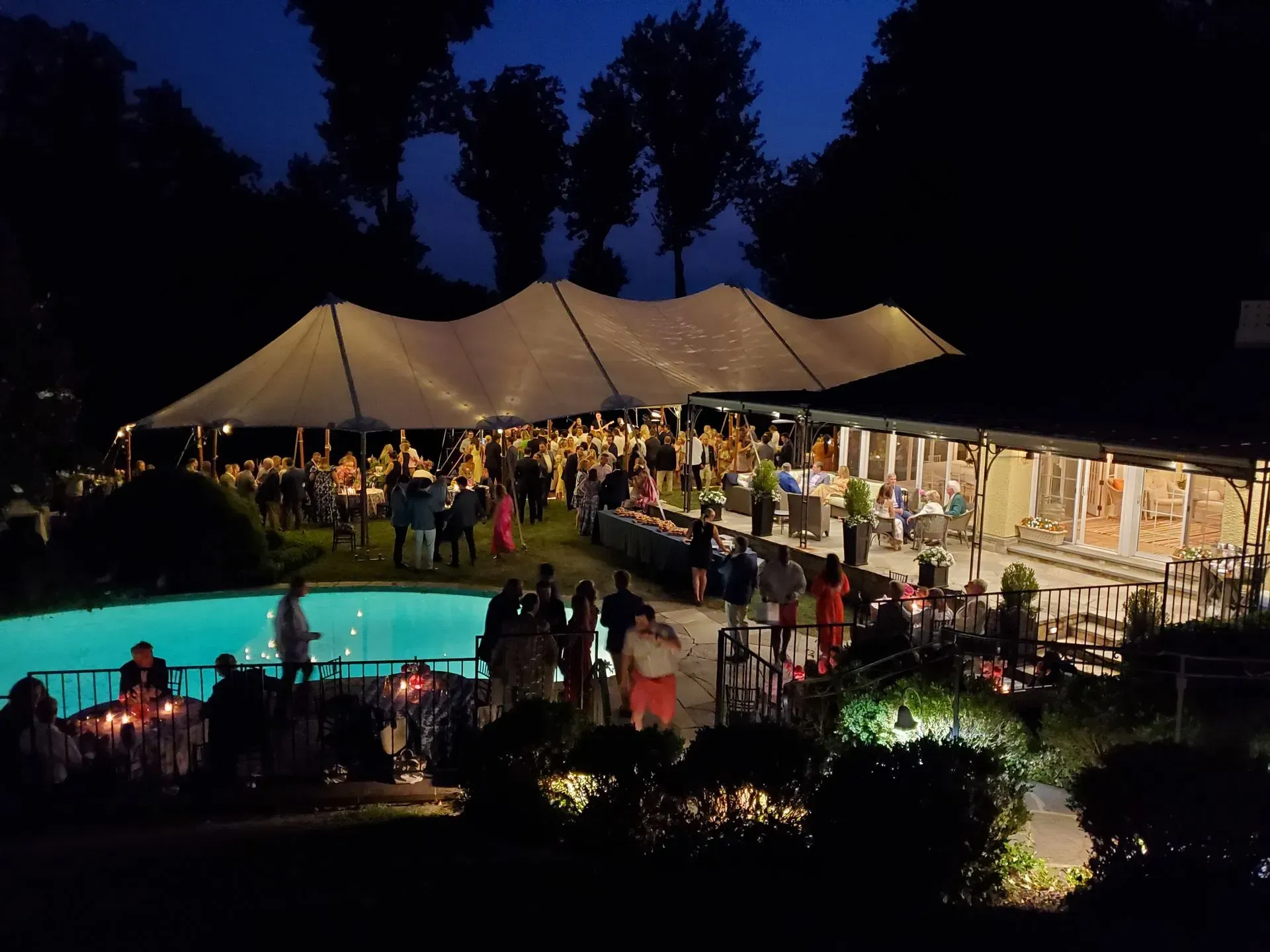 Outdoor evening garden party with a large white canopy tent, swimming pool, and guests dining by tables under lights.