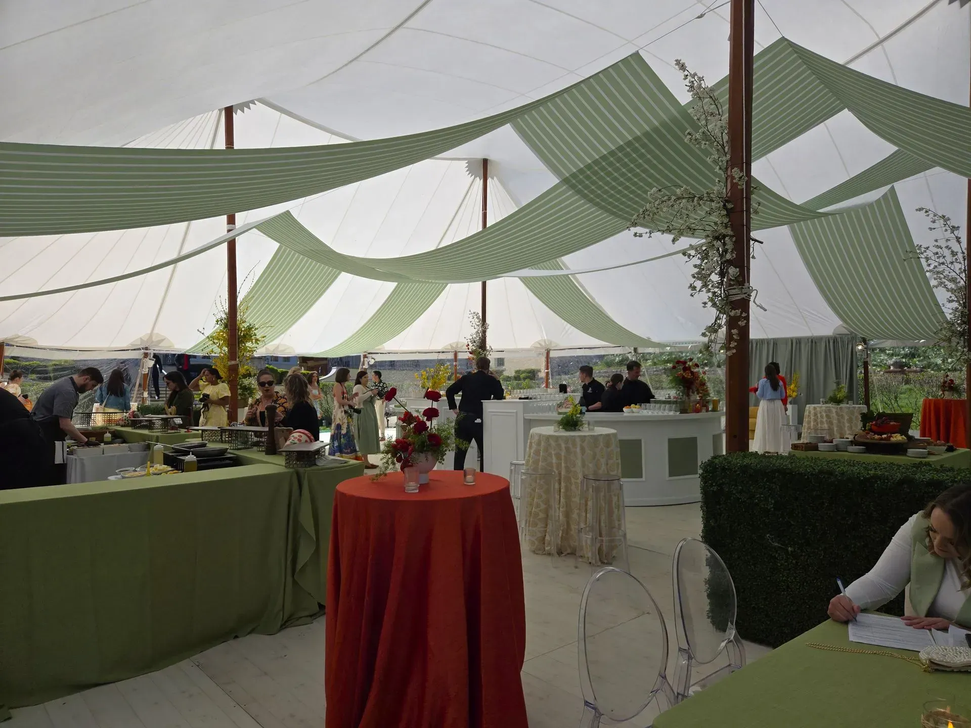 A brightly lit tent with green and white draped fabric overhead, featuring buffet tables, a red cocktail table, and guests.