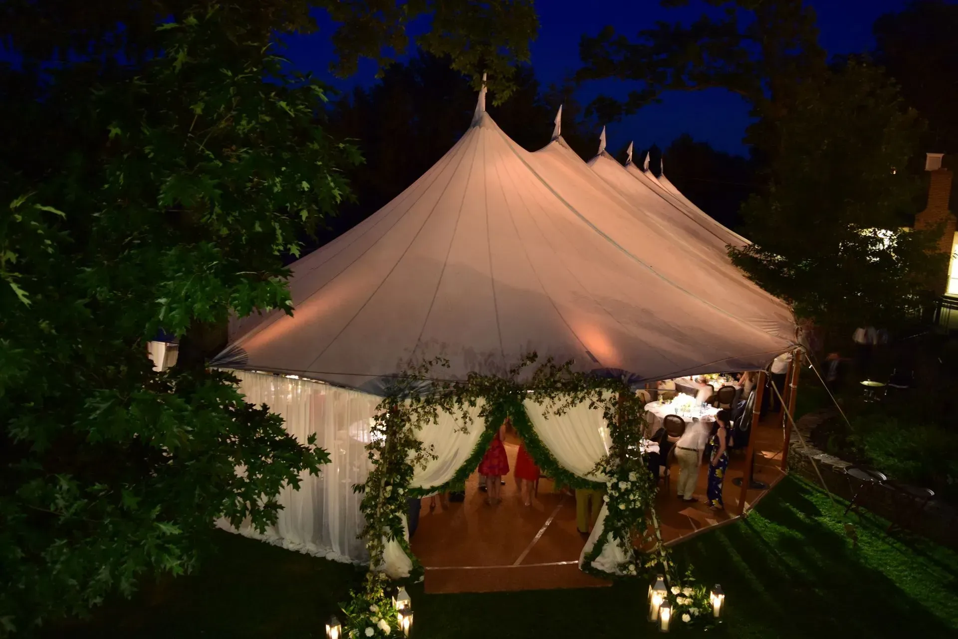 A tall white event tent with arched entrances decorated with greenery stands in a garden at twilight.