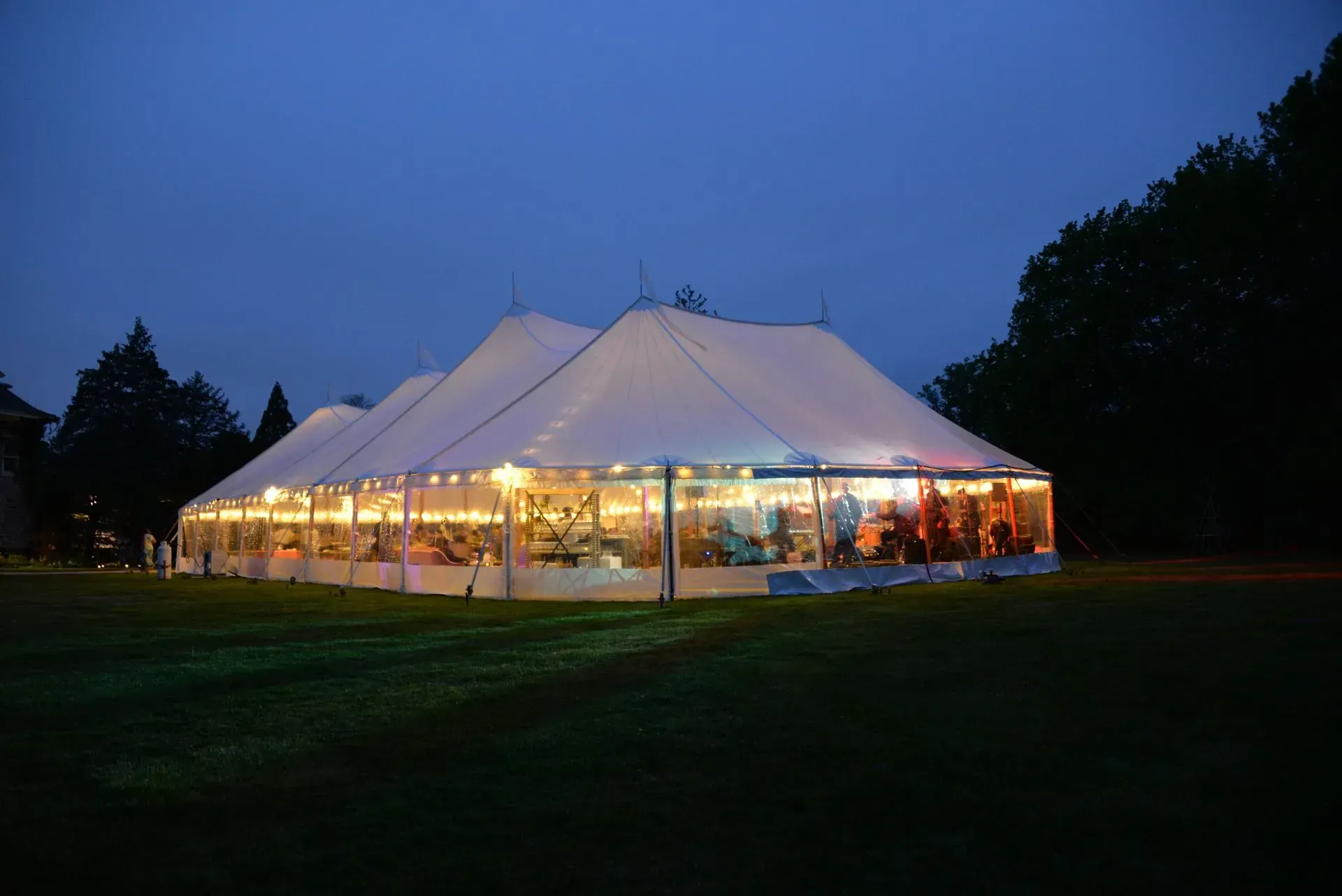 A large, white wedding tent glowing with warm lights at dusk, set in a grassy field surrounded by trees.