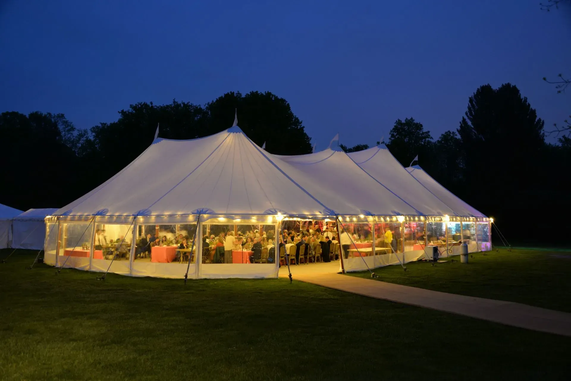 A large, illuminated white event tent glows in a grassy field at twilight, hosting a dinner gathering inside.