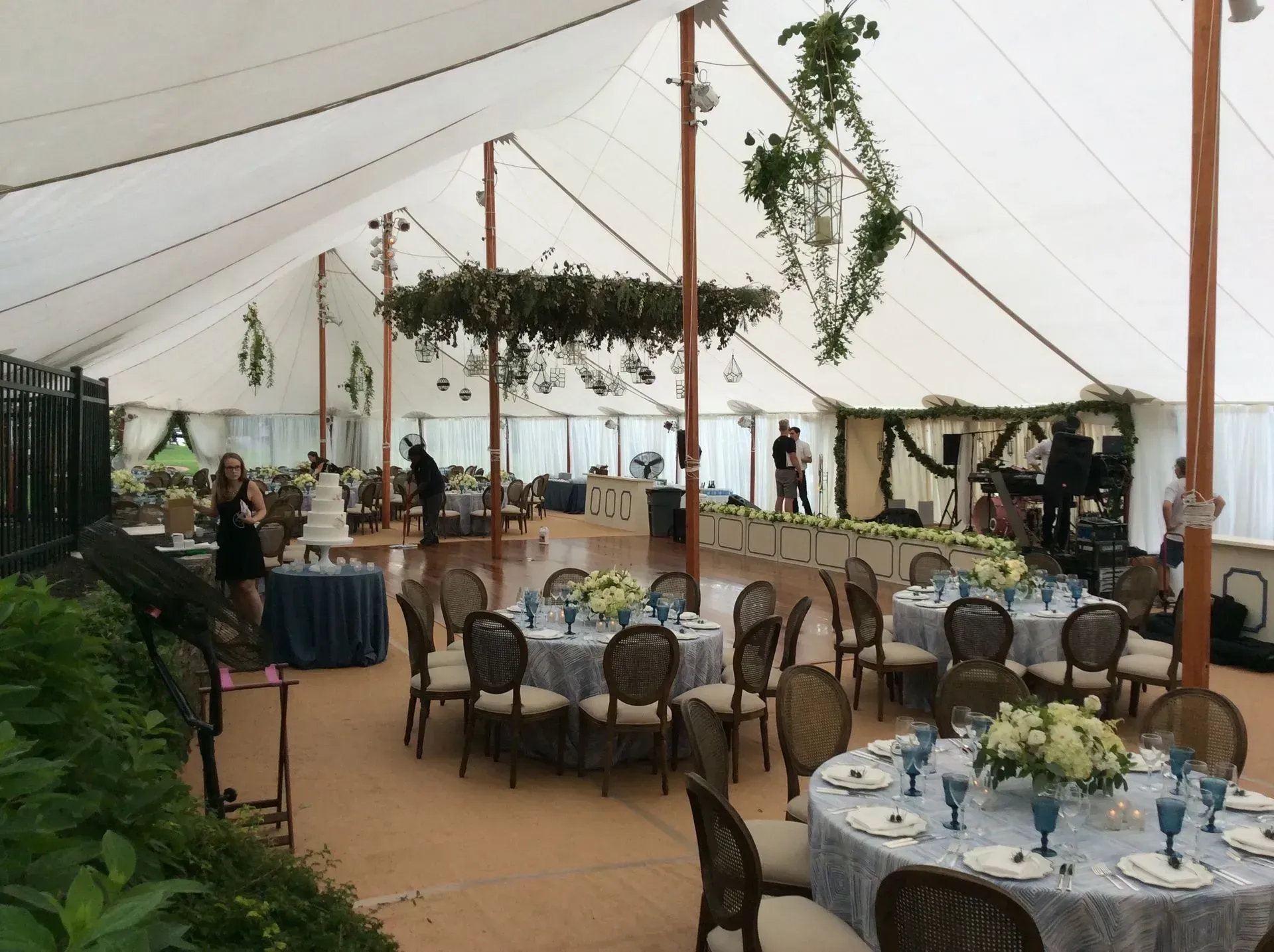 A tented event space featuring round tables with blue linens, floral centerpieces, and hanging greenery decorations.
