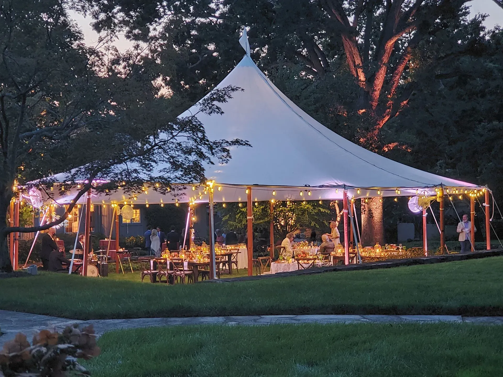 A large white marquee tent lit with string lights at dusk, set on a green lawn surrounded by trees.