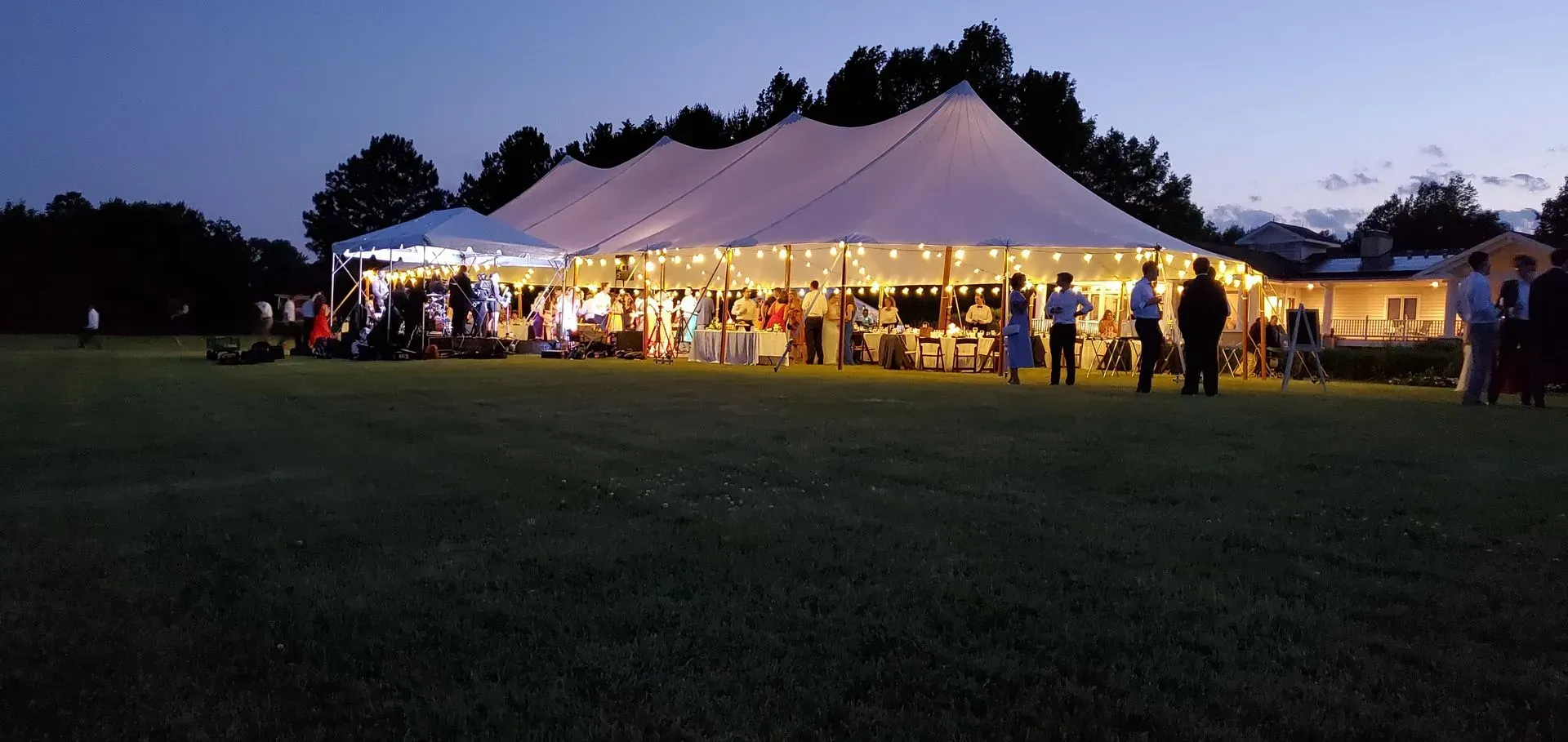 A large, illuminated white tent hosts an evening outdoor event with people mingling on a grassy lawn at dusk.