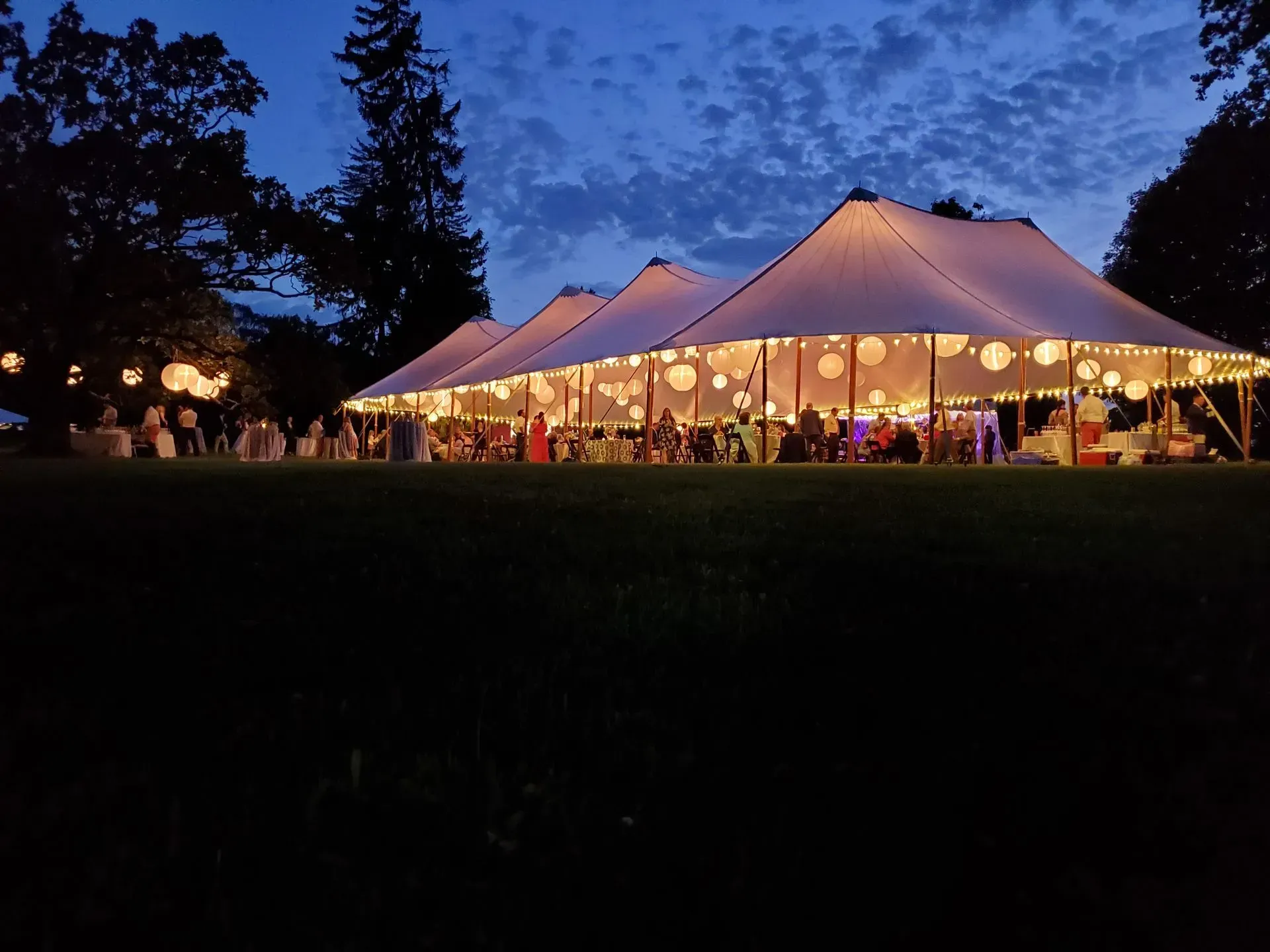 A large, illuminated event tent glows on a grassy lawn under a twilight sky, surrounded by trees with hanging lanterns.
