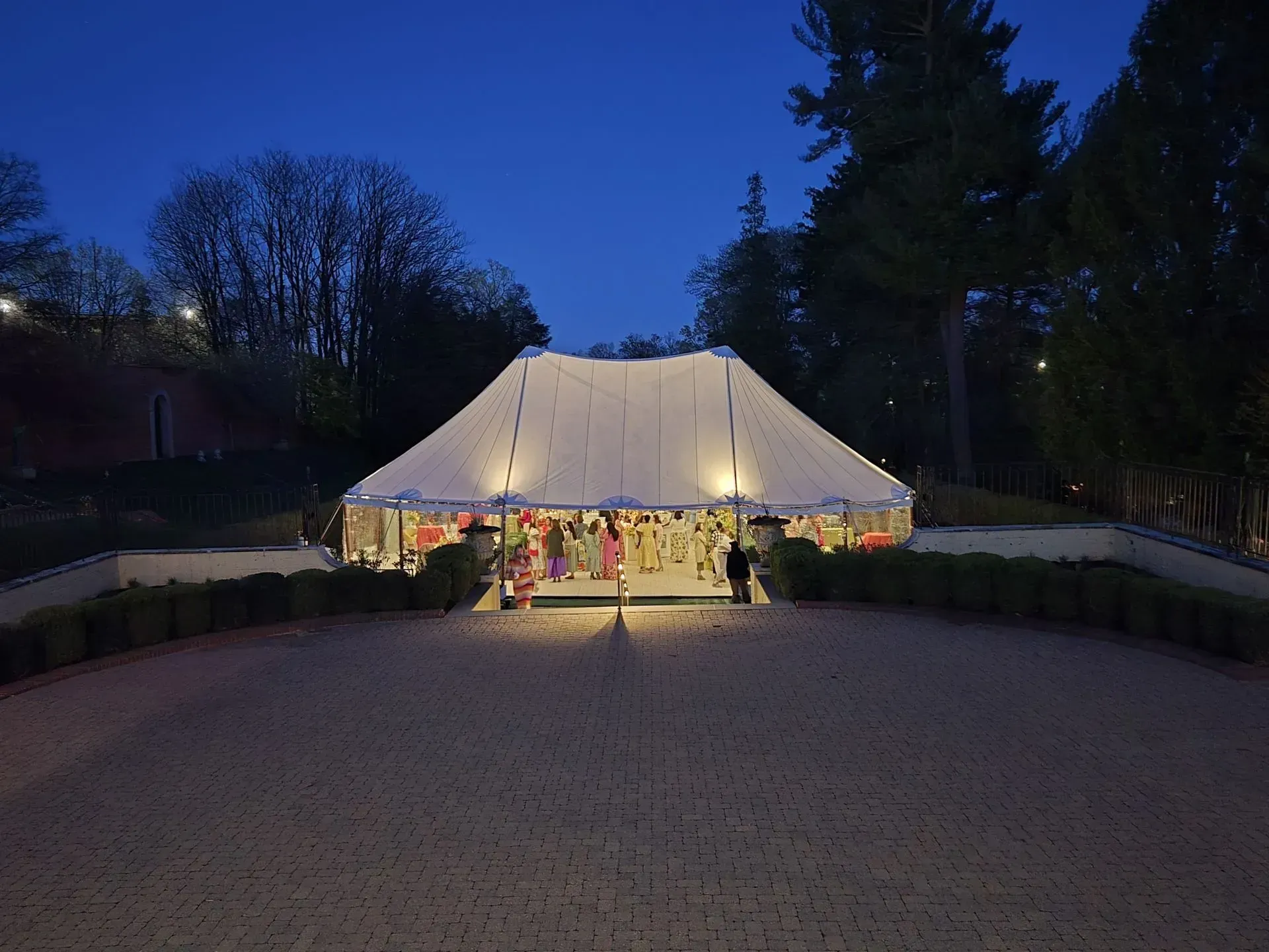 A large, white event tent illuminated at night, situated on a stone patio surrounded by trees and a hedge.