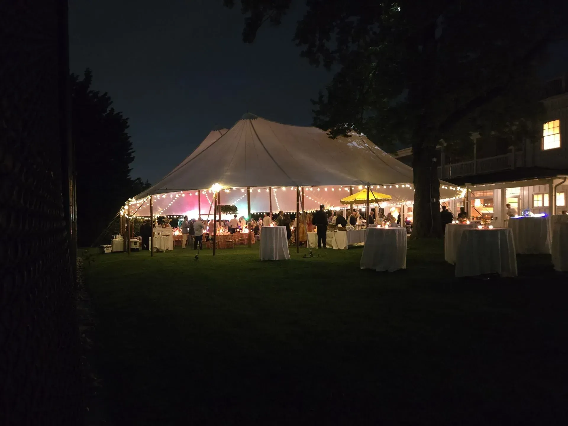 An outdoor evening party under a lit white tent on a lawn with scattered cocktail tables.
