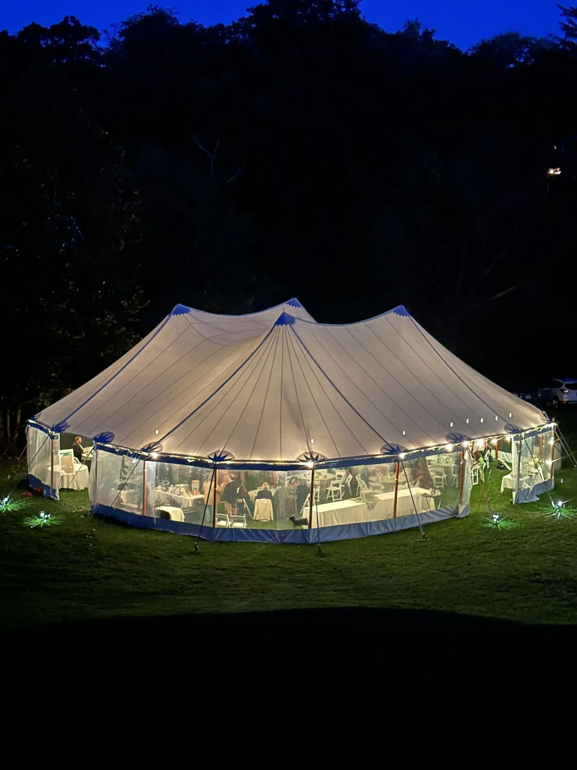 A large, illuminated event tent glows on a dark, grassy lawn at night, revealing tables and chairs inside.