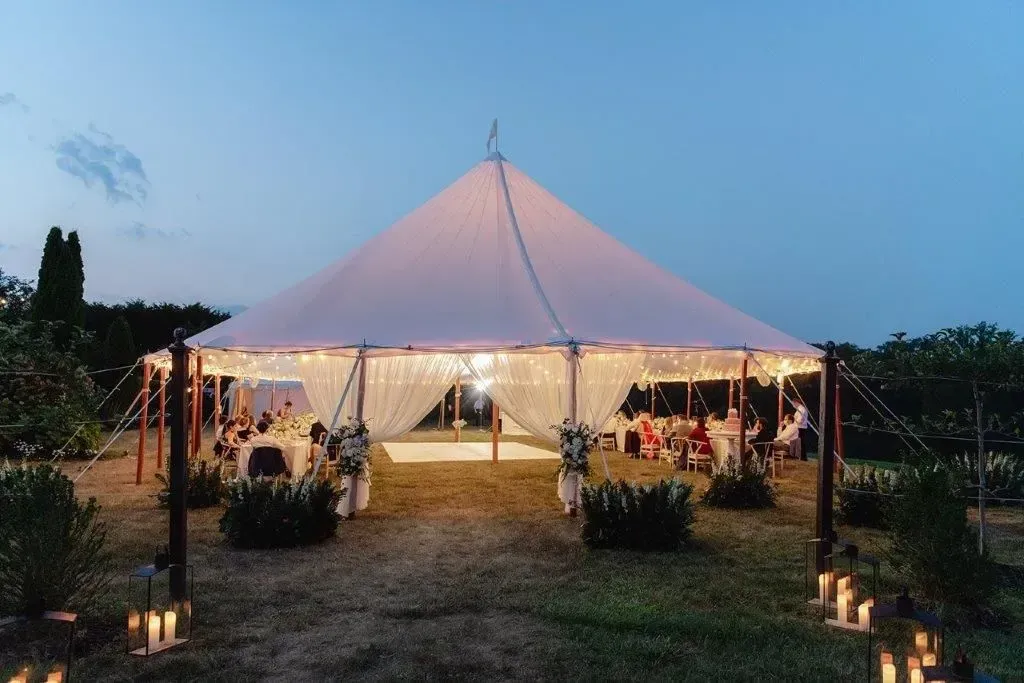 An illuminated event tent sits in a grassy field at dusk, with guests seated at tables inside.