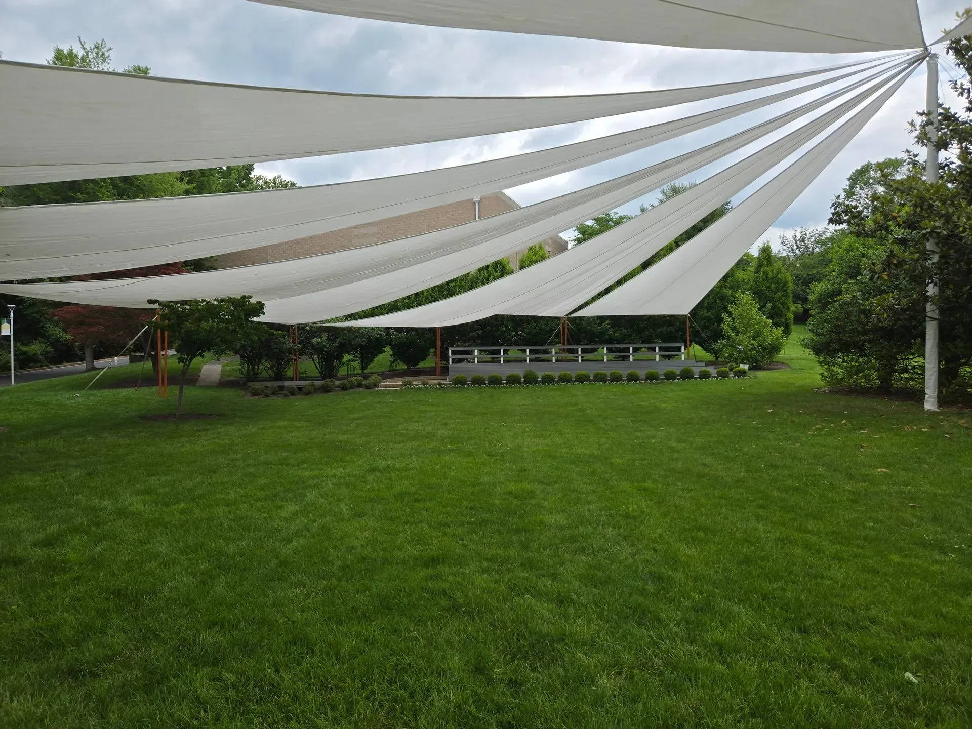 Off-white shade sails stretched over a lush green lawn in a park setting, supported by poles against a cloudy sky.