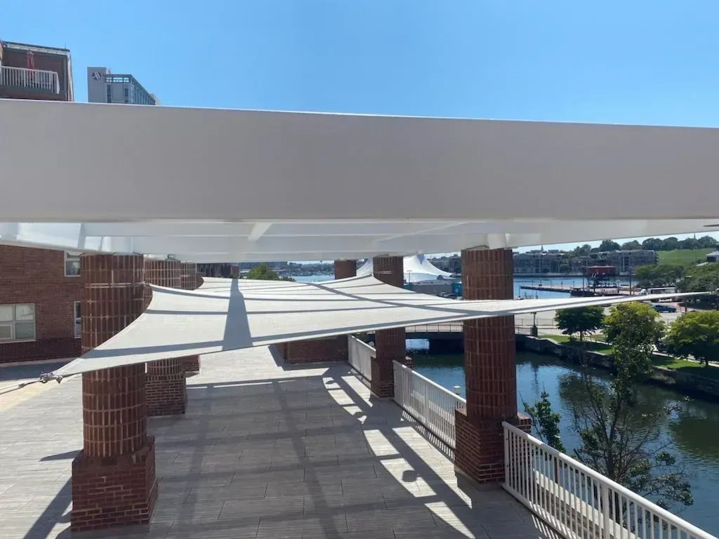 A brick-columned patio with a white overhead shade structure overlooking a calm river on a sunny day.