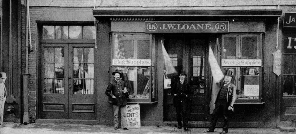 Three people stand in front of the storefront of J.W. Loane, holding signs and fabric flags on a city street.