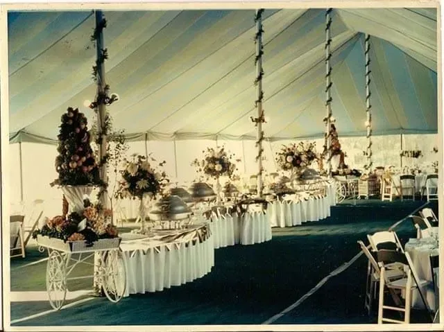 A large event tent with draped ceilings, tables featuring food service equipment, floral arrangements, and dining chairs.