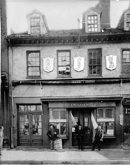 A black-and-white photo of a storefront with three people standing in front and crests hanging on the building facade.