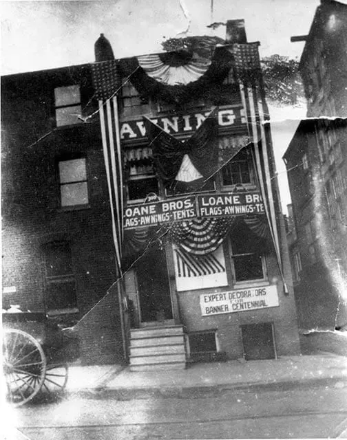 Historic storefront of Loane Bros. decorated with American flags and bunting, with a horse-drawn carriage on the side.