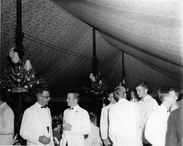 A group of people wearing formal white dinner jackets stand and converse inside a large, draped tent with chandeliers.
