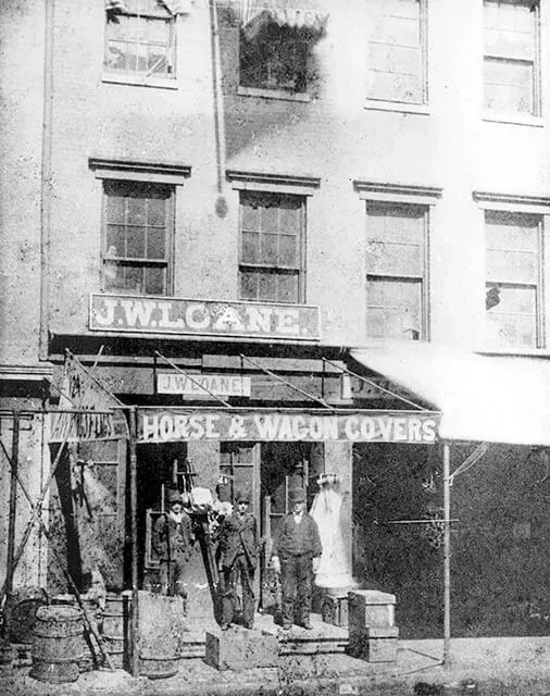 A black-and-white, historic street-level photo of J.W.L. Cane's shop, selling horse and wagon covers, with three men outside.