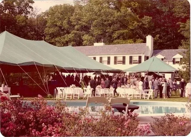 A garden party with green tents set up on a lawn beside a swimming pool, with guests gathered near a large white house.