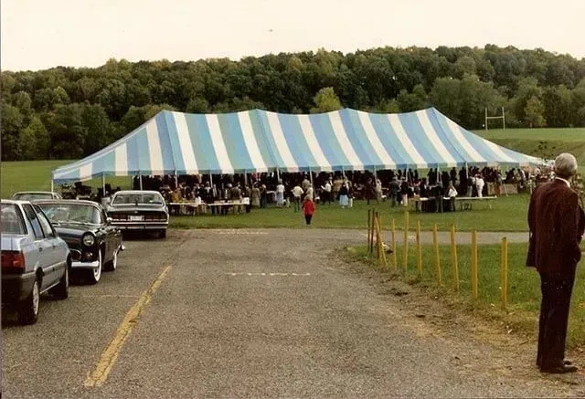 A blue and white striped event tent in a grassy field with people gathered inside, viewed from a parking area.